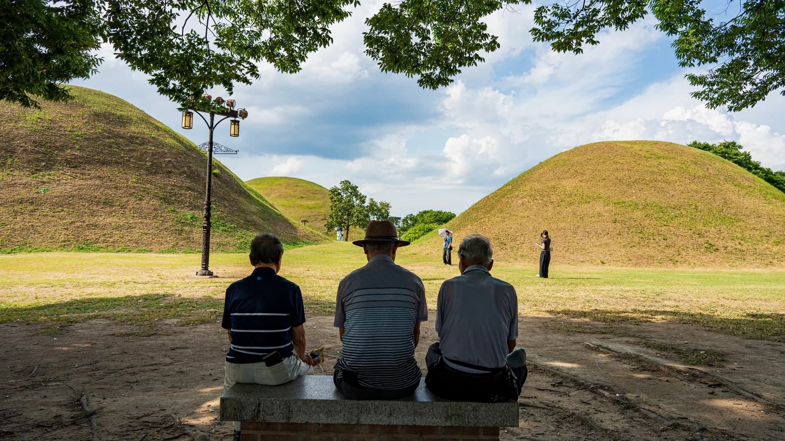 Tre anziani seduti di spalle su una panchina all’ombra davanti ai tumuli erbosi di Gyeongju