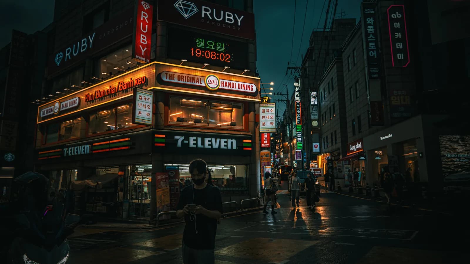 Un 7-Eleven all’angolo di una strada di Seul, tra insegne luminose, passanti e riflessi sul selciato bagnato