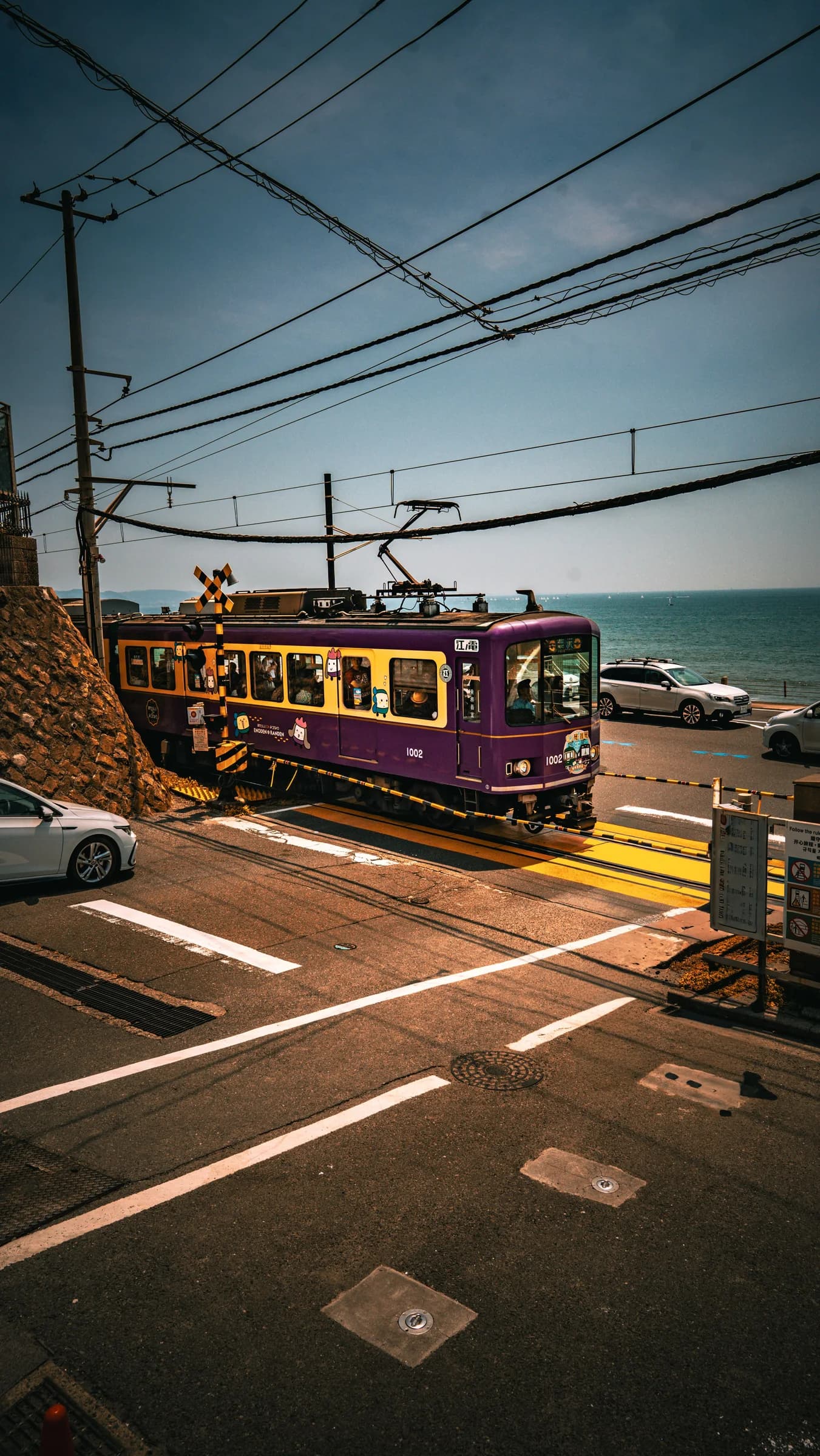 A purple and yellow train crossing a road with the sea behind it