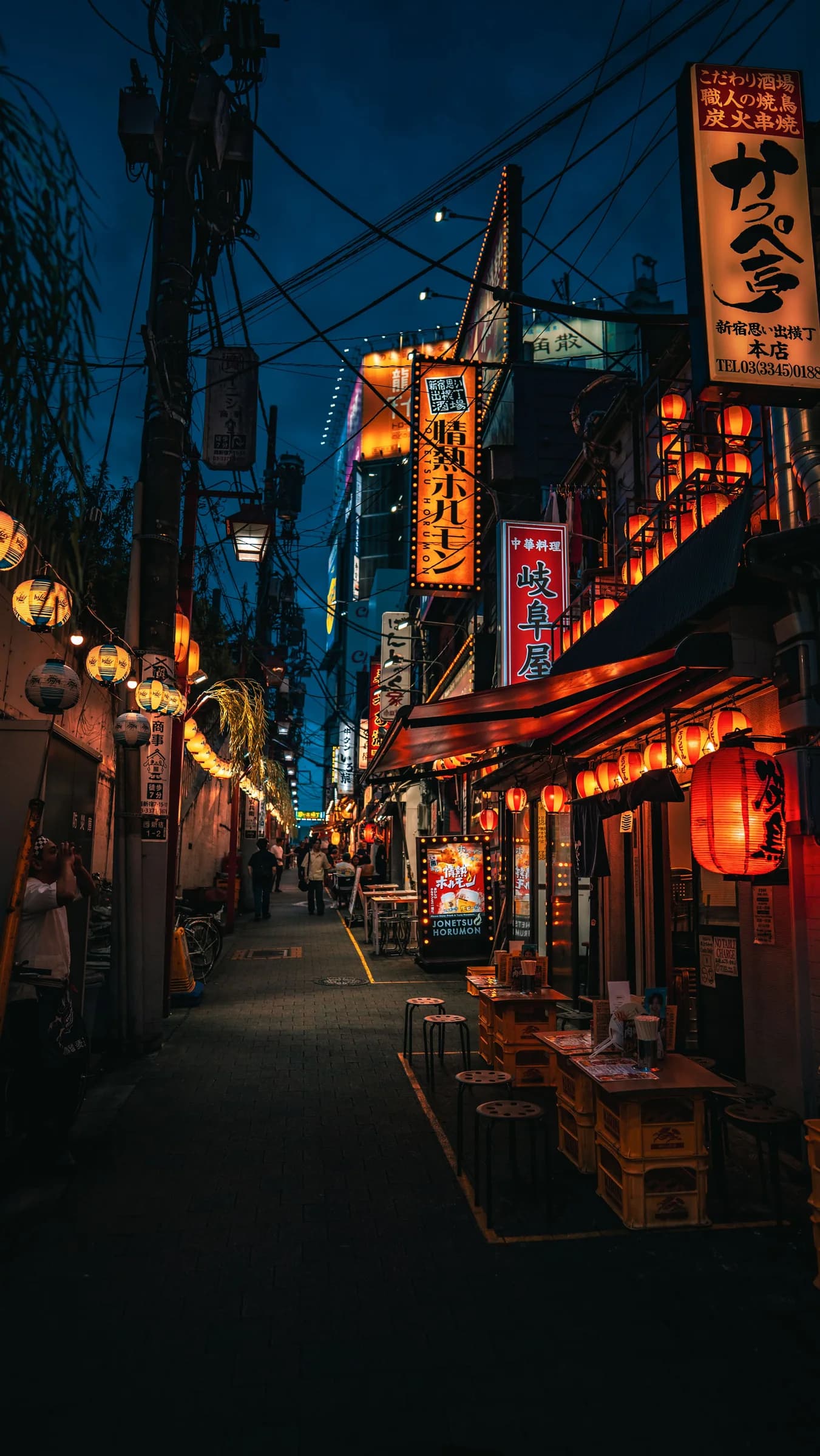 A narrow street at dusk, with orange lanterns and illuminated signs against a blue sky
